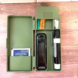 Peut inclure: Un étui de rangement vert olive imprimé en 3D contenant un lecteur de glycémie noir, un autopiqueur, deux lancettes colorées, un petit paquet de bandelettes de test et une boîte blanche avec le texte "DISTRIBUTED BY SAM'S WEST, INC. BENTONVILLE, AR 72716".