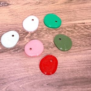 May include: Six flat, irregularly shaped circular tags with a single hole near the top edge. The set includes one white, one light green, one speckled light gray, one light pink, one dark olive green, and one bright red tag, all with a smooth finish.