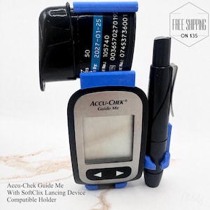 May include: A black and silver Accu-Chek Guide Me blood glucose meter with a blue holder, test strips, and a black lancing device. The text "Accu-Chek Guide Me" is visible on the meter. The holder is compatible with SoftClix Lancing Device.
