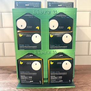 May include: A green 3D-printed display stand holding multiple boxes of Libre 3 Continuous Glucose Monitoring System. Each box is black and yellow, with white sensors and text. The stand has the "Libre 3" logo.