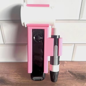 May include: A pink and black device holder with a black device and a pen-like object. The holder is mounted on a white surface with a pink top. The device has a black screen and a circular button.