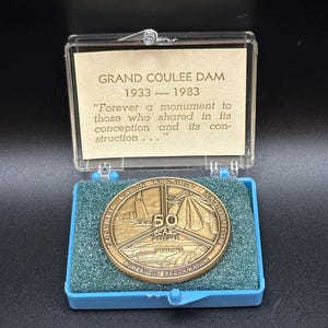 May include: A commemorative coin celebrating the 50th anniversary of the Grand Coulee Dam, 1933-1983, displayed in a blue case. The coin is bronze-colored and features text and imagery related to the dam and the Bureau of Reclamation.