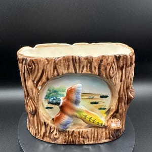 May include: A ceramic planter shaped like a tree trunk, featuring a relief of a flying duck over a marsh scene. The planter has a brown exterior with a light beige interior. The duck has orange, blue, and yellow feathers.