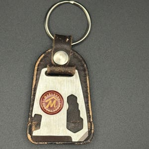 May include: A vintage keychain featuring a multi-tool design with a stainless steel body and a brown leather backing. The keychain has a silver key ring and a circular emblem with the "Marlboro" logo in red and gold.
