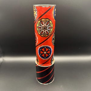 May include: A vintage kaleidoscope with a red body featuring a repeating floral pattern in gold, white, and blue. The base is black with red diagonal stripes. The kaleidoscope is cylindrical.