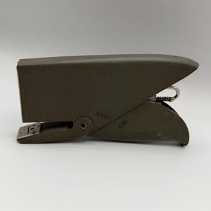 May include: A vintage olive green metal Markwell RX Handi-Clip stapler with a hinged design and a visible metal spring mechanism. The side of the stapler features embossed text that reads Markwell RX Handi-Clip and includes patent information.
