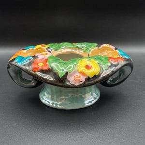 May include: A ceramic flower frog or bowl with a glossy black rim featuring raised, hand-painted flowers in yellow, pink, orange, and blue, set against green leaves. The base is a pale, iridescent light blue with a pedestal design and two handles.