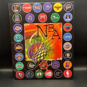 May include: A rectangular black poster featuring the NBA logo in the center with a basketball and net graphic. The border is decorated with circular team logos from various basketball franchises, including the Miami Heat, Bulls, and Lakers.