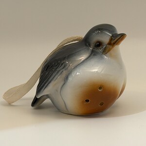 May include: A ceramic bird ornament featuring a glossy finish with a gray head and wings, a white chest, and a brown belly. The bird has three small holes on its front and includes a cream-colored fabric loop for hanging purposes.