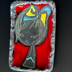 May include: Rectangular enamel art piece featuring a stylized girl with a bow. The girl's face and hair are black with white detailing, set against a vibrant red background. The bow is yellow and blue. The border is textured and gray.
