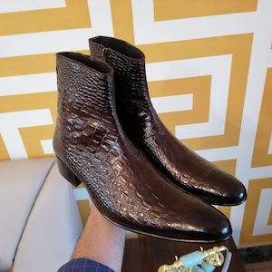 Pode incluir: Um par de botins de couro castanho escuro com um padrão texturizado semelhante ao de um crocodilo. As botas têm bico fino e salto baixo. O fundo apresenta um padrão geométrico amarelo e branco.
