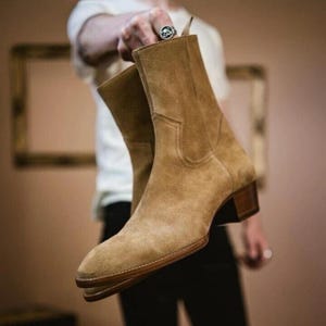 Puede incluir: Un par de botines de ante color canela con tacón grueso. Las botas tienen un diseño occidental clásico con punta afilada y una lengüeta. Las botas miden aproximadamente 20 cm de alto.