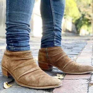 May include: Brown suede ankle boots with a pointed toe and a low heel. The boots have a zipper on the side and are worn with blue jeans. The boots appear to be made of a soft, textured material.