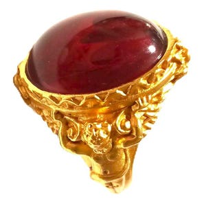 May include: A gold-colored ring featuring a large, oval, deep red gemstone. The ring's band and setting are intricately detailed with ornate carvings, creating a vintage or antique aesthetic. The gemstone is smooth and polished.