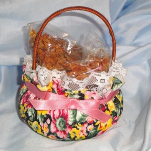 May include: A small fabric basket with a floral pattern in red, yellow, and green on a black background. The basket features a white lace trim, a pink satin ribbon tied in a bow, and a brown handle. Inside, a clear plastic bag holds dried petals.
