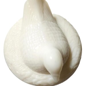 May include: A white ceramic dove figurine, perched on a circular base. The dove is depicted with its head bowed, and its wings are slightly spread. The figurine has a textured surface and a glossy finish.