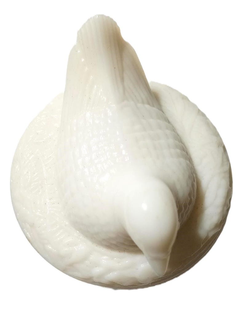 May include: A white ceramic dove figurine, perched on a circular base. The dove is depicted with its head bowed, and its wings are slightly spread. The figurine has a textured surface and a glossy finish.