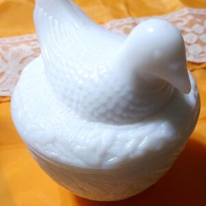 May include: A white, bird-shaped trinket box. The lid forms the bird's body and head, with a textured surface. The base is round and also textured. The box is made of a glossy, opaque white material.