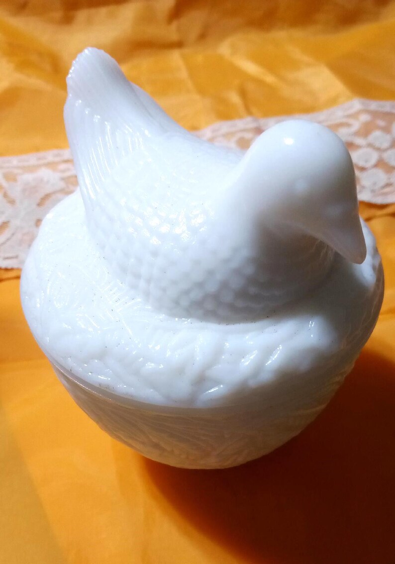 May include: A white, bird-shaped trinket box. The lid forms the bird's body and head, with a textured surface. The base is round and also textured. The box is made of a glossy, opaque white material.
