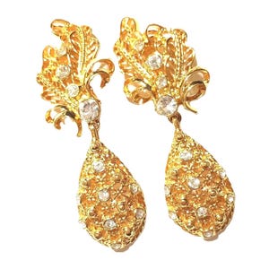 May include: A pair of gold-toned earrings with a leaf-like design, adorned with clear, round crystals. The earrings feature a teardrop-shaped dangle, also embellished with crystals, creating a sparkling, elegant accessory. The earrings are set against a white background.