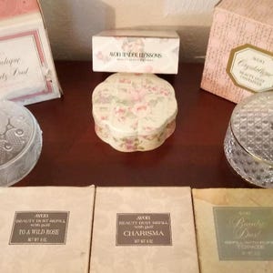 May include: An assortment of vintage Avon beauty products, including decorative glass jars with lids and boxes. The boxes feature text such as "Crystalique Beauty Dust" and "To a Wild Rose". The items are arranged on a dark wooden surface.