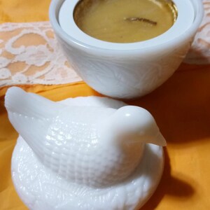 May include: A white, decorative candle holder with a dove-shaped lid. The candle holder has a round base and a white, textured exterior. The candle inside is gold. The dove lid is also white and has a textured surface.