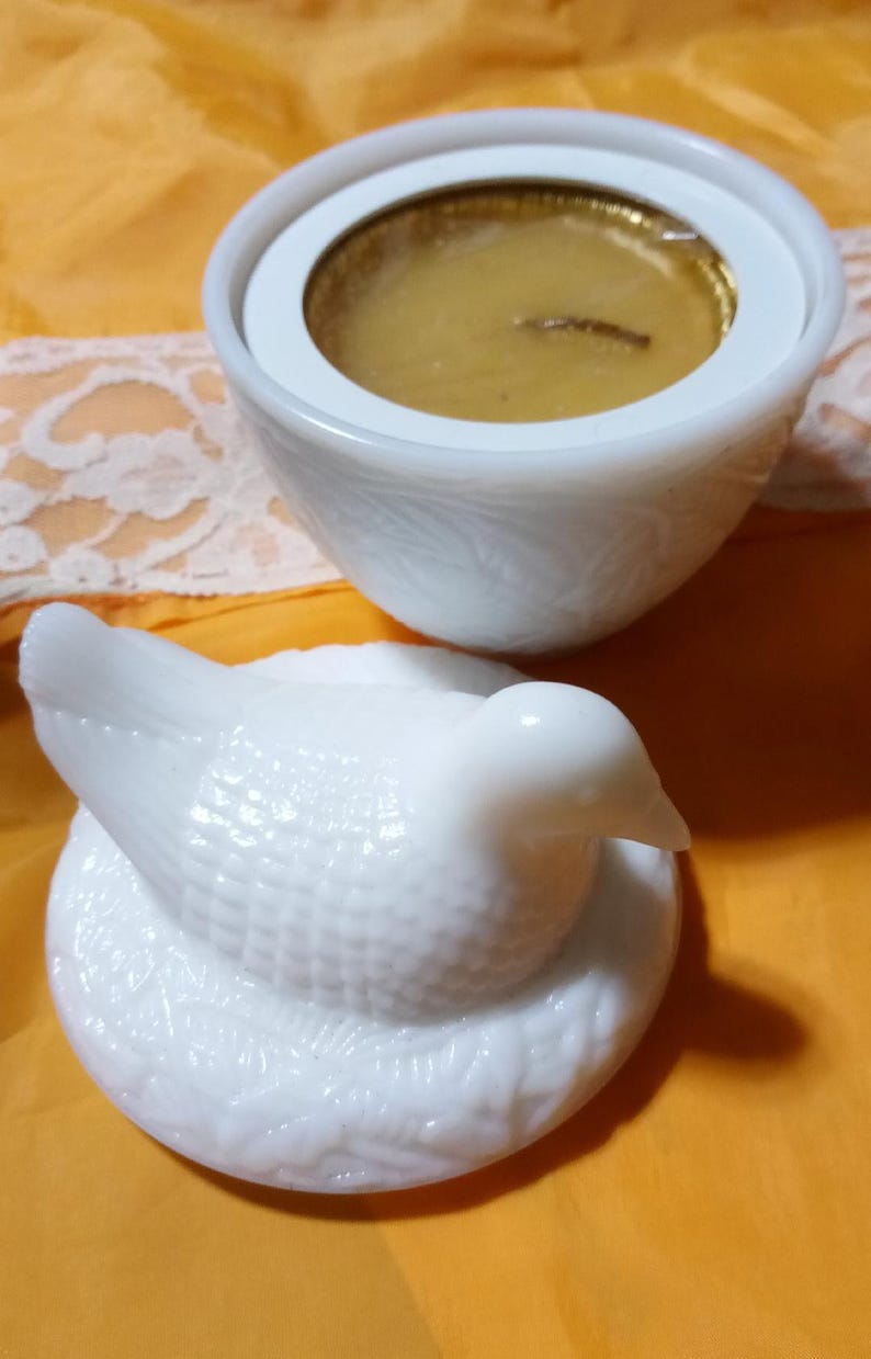 May include: A white, decorative candle holder with a dove-shaped lid. The candle holder has a round base and a white, textured exterior. The candle inside is gold. The dove lid is also white and has a textured surface.