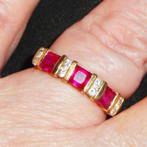 May include: A gold ring featuring alternating rectangular red gemstones and clear, sparkling stones. The ring has a classic design with a gold band and is set with multiple gemstones.