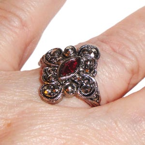 May include: Ornate silver-toned ring featuring a central oval-cut, deep red gemstone. The ring has a cluster design with smaller, round accents and filigree detailing, creating a vintage or antique aesthetic. The ring is on a finger.