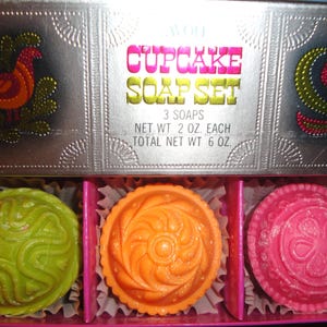 May include: A cupcake soap set with three soaps in a decorative box. The soaps are green, orange, and pink, each with a unique embossed design. The box is silver with floral and bird designs. The text on the box reads "Cupcake Soap Set".