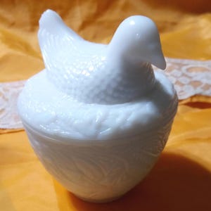 May include: A white, hen-shaped trinket box. The lid is the hen, with a textured body and a smooth head. The base is a rounded bowl with a leaf pattern. The box is made of opaque white glass.