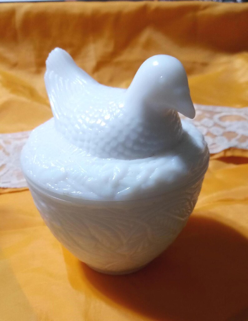 May include: A white, hen-shaped trinket box. The lid is the hen, with a textured body and a smooth head. The base is a rounded bowl with a leaf pattern. The box is made of opaque white glass.