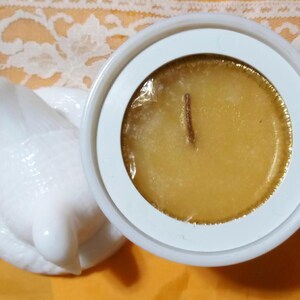 May include: A white ceramic candle holder with a dove-shaped lid. The candle inside is a golden yellow color with a wooden wick. The candle is in a white circular container.