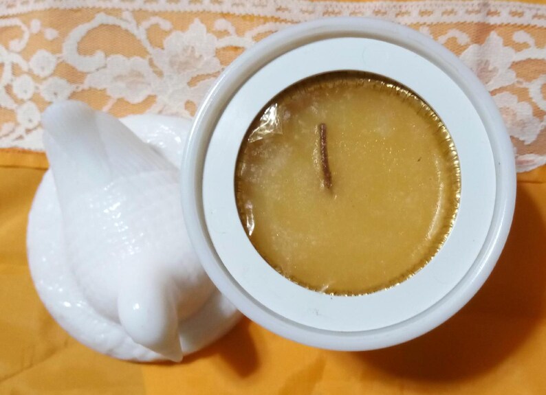 May include: A white ceramic candle holder with a dove-shaped lid. The candle inside is a golden yellow color with a wooden wick. The candle is in a white circular container.