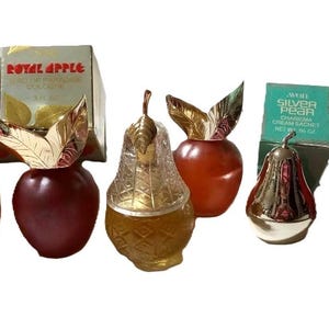 Op de afbeelding: Verzameling vintage parfumflesjes in de vorm van appels en peren, met goudkleurige bladvormige doppen. Twee dozen "Royal Apple" eau de cologne zijn zichtbaar. Eén flesje is peervormig met een goudkleurig, gestructureerd ontwerp.