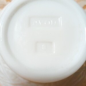 May include: Close-up of the bottom of a white, opaque, vintage Avon container. The base features embossed text reading "AVON" and a smaller rectangular mark below. The container has a textured, possibly floral, pattern on its side.
