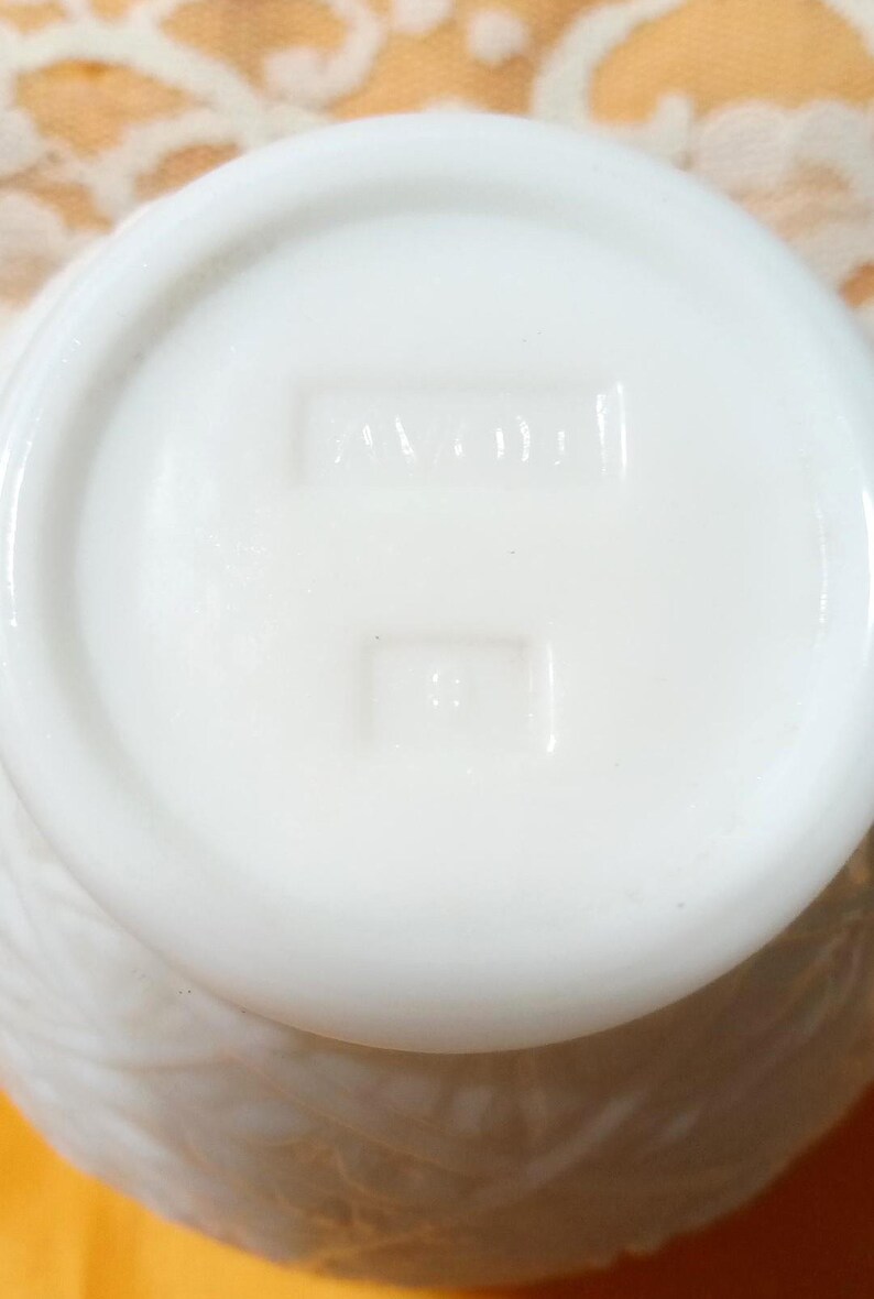 May include: Close-up of the bottom of a white, opaque, vintage Avon container. The base features embossed text reading "AVON" and a smaller rectangular mark below. The container has a textured, possibly floral, pattern on its side.