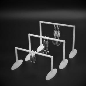 Op de afbeelding: Drie witte oorbellen display stands van verschillende hoogtes, elk met een horizontale balk en ovale basis. Verschillende paar oorbellen worden op de balken getoond. De achtergrond is effen zwart.