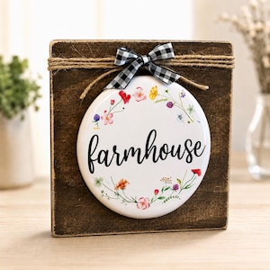 May include: A farmhouse-themed decorative sign. The sign features a white, circular design with the word "farmhouse" in black script, surrounded by a floral wreath. It is set against a dark brown wooden block, adorned with twine and a black and white checkered bow.