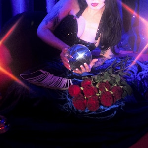 May include: A woman with long black hair and red lipstick holds a crystal ball. Red roses and tarot cards are in front of her. The scene is lit with candles and a blue backdrop.