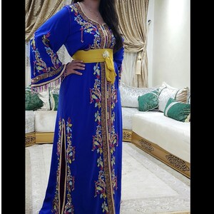 May include: A royal blue caftan with intricate gold embroidery and a yellow sash. The long-sleeved dress features a high slit and ornate detailing along the neckline and sleeves. The caftan is a traditional garment.