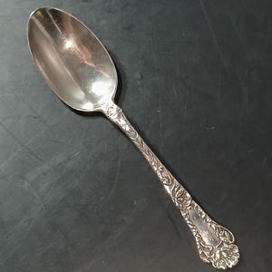 Puede incluir: Una cuchara de plata con un cuenco ovalado y un mango decorativo. El mango presenta un diseño floral ornamentado. La cuchara probablemente está hecha de plata de ley o un metal similar. La cuchara es una pieza de cubertería, adecuada para servir alimentos.