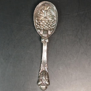 May include: A silver spoon with a detailed pineapple design on the bowl. The handle features ornate detailing and a decorative bow. The spoon is likely made of silver or a silver-plated metal.