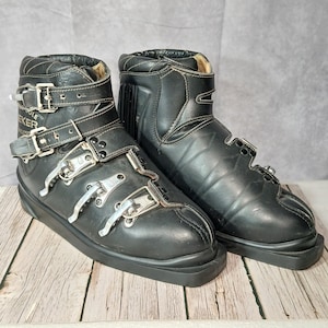 Pode incluir: Par de botas de esqui em couro preto com várias fivelas e correias de metal prateado. As botas têm bico arredondado e sola grossa. As botas estão sobre uma superfície de madeira.