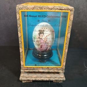 Puede incluir: Una exhibición enmarcada con un huevo decorativo con una imagen pintada de una mujer y un menú para la 4ª Conferencia Anual KILICO 1982. El menú incluye platos como ensalada de pomelo y camarones y sopa clara de rabo de buey.