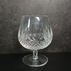 May include: A clear, stemmed brandy snifter with a rounded bowl and a diamond-cut pattern on the lower half. The glass sits on a circular base and has a short stem. The background is a dark gray.