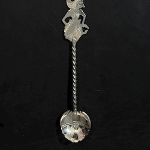 May include: A silver spoon with a detailed design. The spoon features a figure at the top, a twisted stem, and a detailed bowl. The spoon is likely a decorative or collectible item.