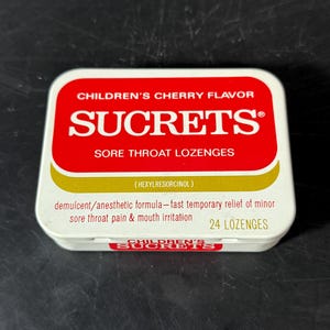 Op de afbeelding: Een rechthoekig wit metalen blikje met Sucrets Children's Cherry Flavor keeltabletten. Het deksel heeft een rood paneel met witte tekst, een gouden gebogen accent en de tekst: (Hexylresorcinol) demulcent/anesthetic formula 24 lozenges.