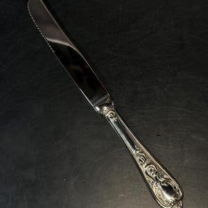May include: A silver-toned dinner knife featuring a serrated blade and a handle decorated with intricate embossed scrollwork and floral patterns. The metal surface is polished and reflective, showing a detailed design along the entire handle.