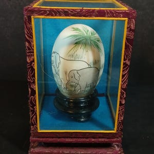 Puede incluir: Un objeto decorativo en forma de huevo con una escena pintada de elefantes y palmeras, exhibido dentro de una vitrina con un marco de terciopelo granate y un interior azul. El huevo descansa sobre un soporte negro.
